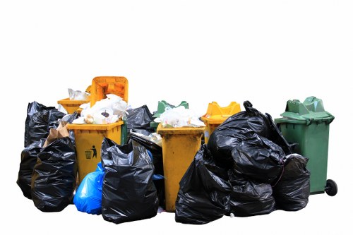 The image displays a collection of various waste disposal bins placed outdoors on a paved surface, likely a driveway or designated rubbish area. There are seven bins in total, including a yellow bin positioned centrally with a slightly open lid revealing white rubbish inside, a smaller green bin with a closed lid on the right, and an orange bin with a closed lid behind the yellow one. Additionally, there is a blue bag leaning against the yellow bin on the left and multiple black rubbish bags piled around the bins, some partially open showing mixed waste. The black bags appear to be filled, with creases and crumples on the surfaces, indicating they are tightly packed. The green and yellow bins have a smooth plastic finish, typical of outdoor waste containers, while the black bags are made of durable plastic material. The scene is well-lit with natural daylight, and no background scenery is visible, focusing solely on the waste items, which are typical of rubbish removal services carried out by companies like Commercial Waste Ickenham, in the local area near Ickenham or the UB10 postcode district.
