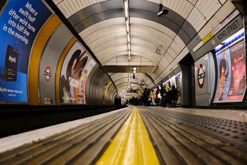 A photograph of a modern underground metro station tunnel showing a curved platform with a black tunnel entrance in the background. The platform surface is smooth and dark, with a yellow tactile pavin