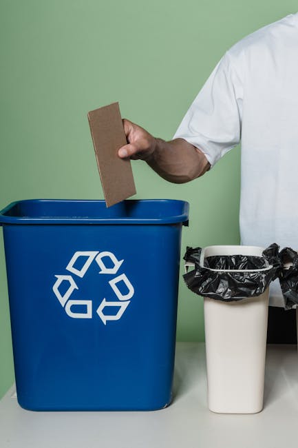 A person wearing a white shirt is holding a rectangular piece of cardboard in their right hand, positioned above a blue recycling bin that features a white recycling symbol on the front. To the right 